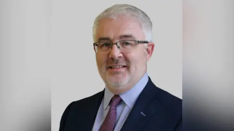 PA Media Michael Bloomfield has white hair, brown eyebrows, and white stubble. He wears half rimmed glasses and a navy suit, with a stripy shirt and pink tie