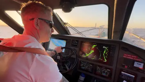 BBC A coxswain wearing sunglasses and a white, short-sleeved shirt sits in the cabin of a fast boat with his hand on a steering wheel. Computer screens on a dashboard show navigation details. Through the front window a wide river can be seen, with the tall Grimsby Dock tower in the distance. The sky is blue and orange as the sun sets.