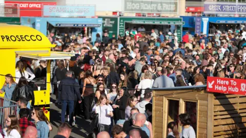 Tees Valley Combined Authority Crowds of people at a food market in Middlesbrough. Food vans surround the square selling various food. A yellow van to the left advertises prosecco. Three vans to the back of the crowd are selling scampi and chips, hog roasts and Greek street food.
