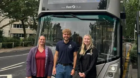 UEA Two women and a man standing in front of a grey Norwich electric First Bus