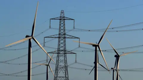 PA Media A group of five wind turbine, in shadow against a blue sky, stand in front of a large electricity pylon with cables stretching to either side.