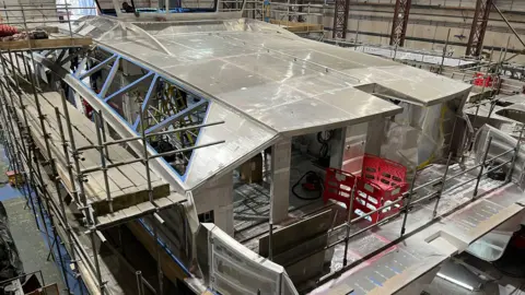 BBC A metallic shell that will come to be the new ferry. It is rather large and there is lots of scaffolding up around it.