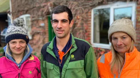 Luke Deal/BBC A man stands in the middle of two women. He is wearing a green farming overall and has dark short hair. The woman to his left is wearing a pink coat and wooly hat. The woman to his right is wearing an orange coat and cream wooly hat.