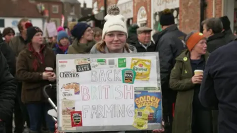 BBC/Spencer Stokes A young woman holds up a sign that reads "back British farming". Several logos of well-known brands are glued to the sign such as Heinz, Cheerios and Quavers crisps. She stands in the middle of the market square, surrounded by others who joined the rally.