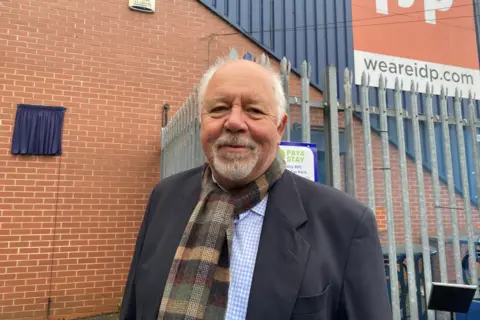 Peter Walters wearing a black blazer and white and blue checked shirt with a brown, green and being scarf.