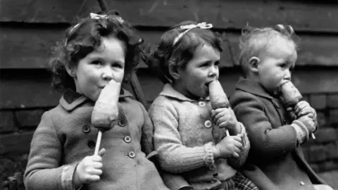 Getty Images Black-and-white photo of three children sitting on a bench eating some kind of vegetable, possibly carrots, on a stick.