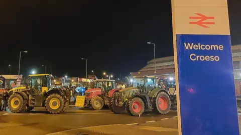 Dyfan Eilir Jones About a dozen large tractors block a road next to Holyhead port. the welcome sign with Irish Ferries and Stena Line names and logos is in the foreground