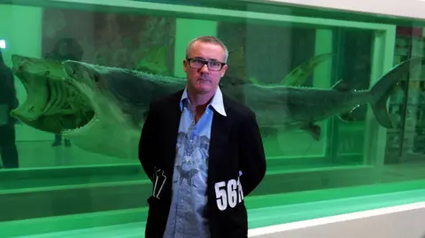 PA Media A man in a baggy blue shirt and black jacket with numbers on the side stands in front of a piece of artwork made of a shark, cut lengthways and suspended in formaldehyde.