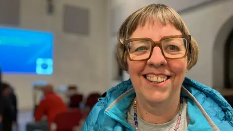 A woman with glasses and a puffer in focus, with a meeting and presentation in the background.