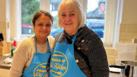 Two women standing together in a kitchen. They are wearing blue aprons with "Friends of Di's Kitchen" written on. Sanju on the left has long dark hair in a pony tail and is wearing a beige jumper and Sharon has grey hair and is wearing a dark top.