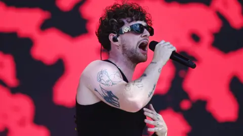 BBC Tom Grennan singing into a microphone while performing live on stage at Glastonbury. He has short brown, curly hair and is wearing sunglasses, a blank tank top and has tattoos on his right arm and left knuckles.