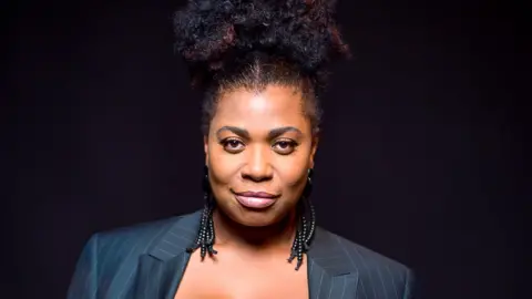 ATG Entertainment Brenda Edwards is dressed in a black pinstripe suit with long black earrings coming from her ears