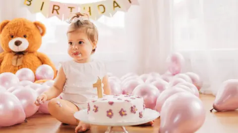 Getty Images A baby girl sits cross-legged on the floor in front of her first birthday cake decorated with purple flowers and a one-shaped candle. She's surrounded by pink balloons, a giant teddy bears sits in the background.  
