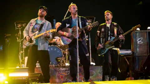 BBC Coldplay on stage with Chris Martin up front and a lead guitarist to the left and bass player to the right. There is also a drummer in the background and yellow lighting. 