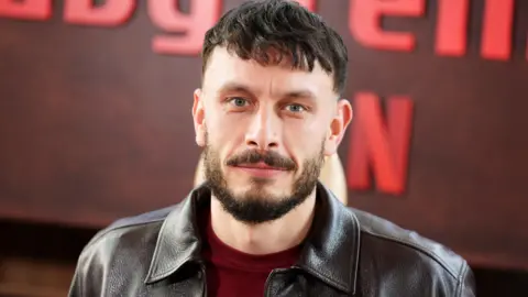 Getty Images Actor Richard Gadd in a close-up wearing a burgundy sweater and leather jacket, standing in front of a Baby Reindeer sign at the series launch