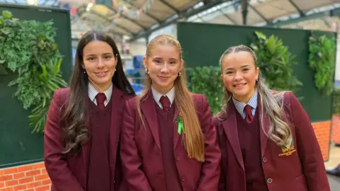 Three girls wearing burgundy school uniforms including a tie, jumper and blazer. Two have blonde hair one has brown.