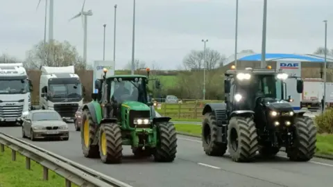 Mick Moore Two tractors drive down a dual carriageway as traffic starts to build up behind them. 