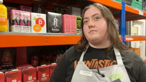 BBC A woman stands in front of drinks products on a supermarket shelf. She has straight brown hair and is wearing a black top and a grey apron. The apron has a green "Your Co-op" logo on it. 