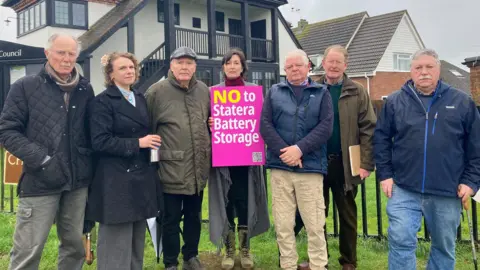 Katharine Da Costa/BBC Seven people stand in a row outside a council office, in protest against the proposed battery site. A lady in the middle holds a bright pink sign saying "NO to Stater Battery Storage". There are two women and five men, all wearing winter coats.