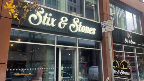 BBC An image of Stix and Stones, a glass storefront has a black sign with white font reading "Stixs and Stones."
