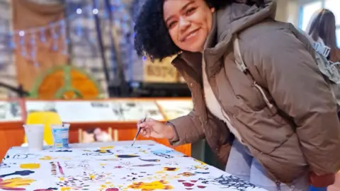 Bradford Council A woman with dark hair and waring a grey coat, jumper, jeans and a backpack is holding a paintbrush. She is leaning over a cloth on a table. The cloth is decorated with patterns and pictures in bright colours.