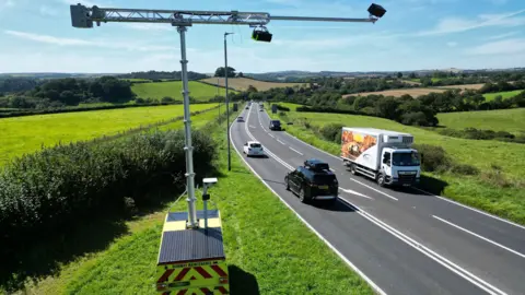 Vison Zero South West Two cameras are suspended from a high metal gantry that is based on the side of the road but arcs out above and across it. It is daytime and there are fields either side of the three-carriage road which has a lorry and three cars in the foreground.