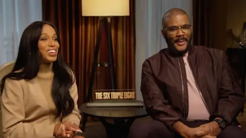 Two people sit on chairs with a table in the middle that has a little sign reading 'The Six Triple Eight' in capitals. Behind them are brown curtains and a tall floor lamp with a white shade. On the left sits a woman with long black straight hair. She is wearing a caramel coloured jumper and she has her hands clasped together, and is smiling. The man next to her has a shaved head, black beard and moustache and wears silver framed glasses. He is wearing a dark coloured bomber jacket over a light coloured shirt.