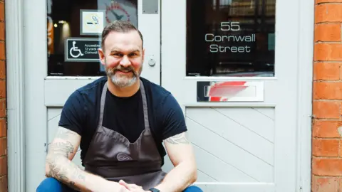 Claire Lishman PR Glynn Purnell in a black t-shirt and apron, sat outside a white door at the front of his restaurant in Birmingham