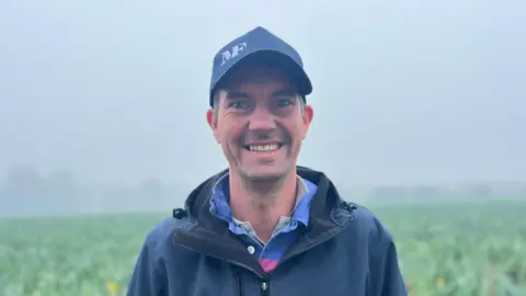 BBC Phil Le Maistre is a man with short hair. He is wearing a navy baseball cap with a blue and pink collared shirt, underneath a navy waterproof. The background is an out-of-focus misty field.