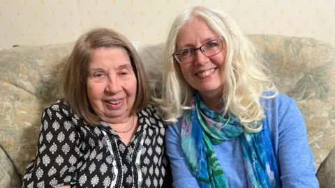 BBC Mother and daughter, Maggie and Lucy are sitting, smiling on a sofa. Maggie is wearing a black and white top and Lucy is wearing a pale blue jumper with a blue, pink and green scarf.