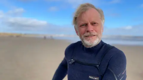 A middle-aged man wearing a blue wetsuit stands on a sandy beach under a blue sky. He has fair hair, which is blowing in the wind, and a white beard