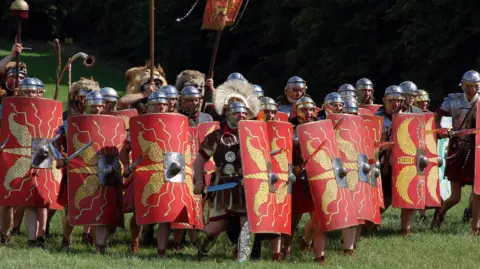Ermine Street Guard Modern Roman military reenactors, holding large rectangular red shields and wearing polished metal armour, form a line as if preparing for battle