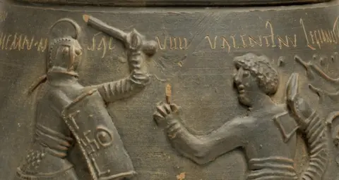 Colchester and Ipswich A relief sculpture on a ceramic cremation vessel decorated with a gladiator fight between a secutor and retiarius, Colchester, 2nd century AD.
