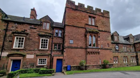 BBC The Prior's Tower by Carlisle Cathedral. The three-storey structure is red sandstone and has windows on the first and second floors. It is flanked by the other buildings also made of sandstone.