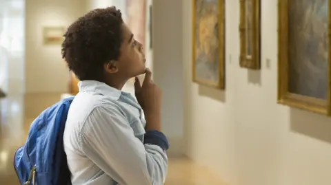 Getty Images A boy in a light blue shirt and carrying a blue bag looks at a collection of paintings hanging on a wall.