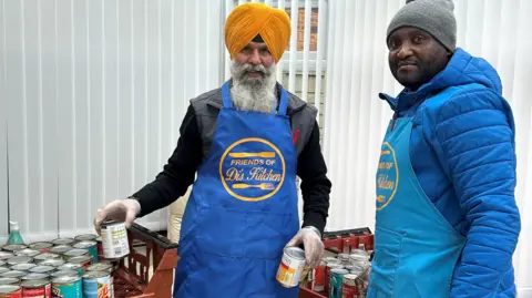 A man wearing a turban with a grey beard holds tins of food in his hands. Another man wearing a hat stands next to him. Both are wearing blue "Friends of Di's Kitchen" aprons.