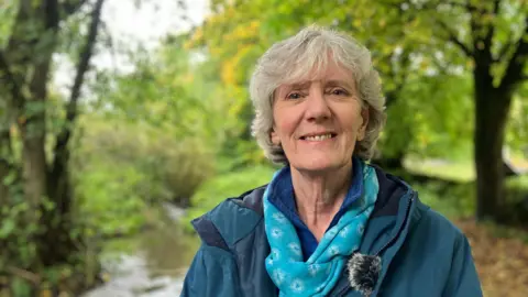 A woman in focus, in front of Tideswell Brook, a small stream in a leafy forest in the Peak District.