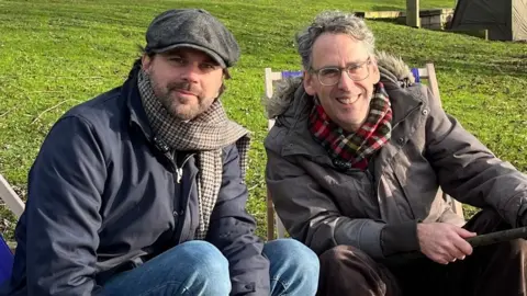 BBC/Lily-May Symonds BBC Reporter Justin Dealey is sat next to Robert Wilfort on a pair of deck chairs. Both are wearing coats and scarfs. Robert is holding a fishing rod.