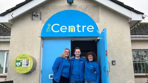 BBC Rebecca Woods, Mitch Hill and Sharon Barnes stand in front of the entrance to The Centre. They are all wearing blue tops with The Centre branding. The door of the building is blue with the The Centre logo on the top.