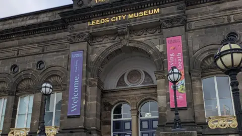 BBC The entrance to Leeds City Museum - an old stone building with its name emblazoned in gold above the doors, which are glassed and blue.