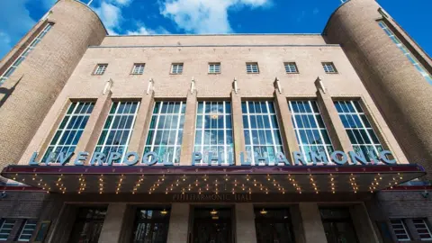 Royal Liverpool Philharmonic Fawn brick building with engraved glass of 3-storey height with receding tiers of parapets. Symmetrical front: between semi-circular stair turrets and a canopied entrance are 7 large vertical metal windows separated by piers topped by jagged abstract motifs.