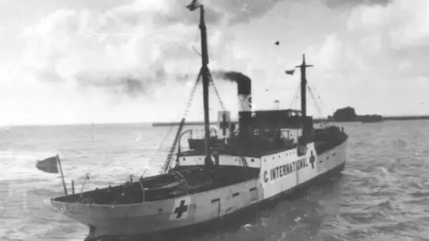Priaulx LIbrary The SS Vega ship in the water in Guernsey's Saint Peter Port harbour in 1944. The white ship has two foldable masts with the red cross symbol painted on it in three locations.