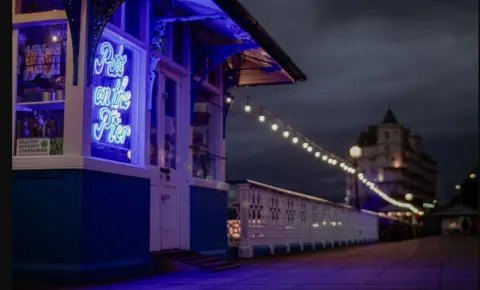 Llandudno pier at sunrise with a sign lit up in a blue shop window and the railings lit by overhead string lights