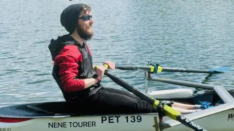 Camsight Alex Innes rowing in a boat. He is wearing glasses, a black hat, a black and red coat, and has both of his hands on oars. He has brown hair and a beard and is smiling. The boat is white and has "Nene Tourer" written on the side.