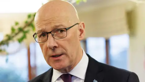 PA Media John Swinney, a bald man with glasses wearing a darks suit and white shirt, looks towards the camera in a close-up shot 
