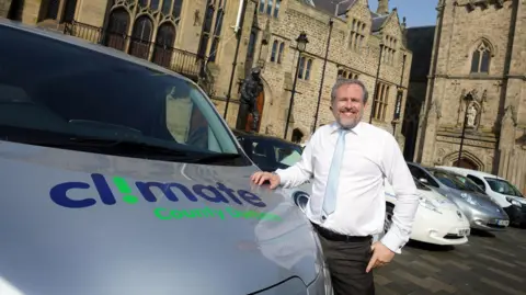 Durham County Council Councillor Mark Wilkes. He is wearing a white shirt and light blue tie. He is standing next to a council van which has the words Climate County Durham printed on its bonnet.