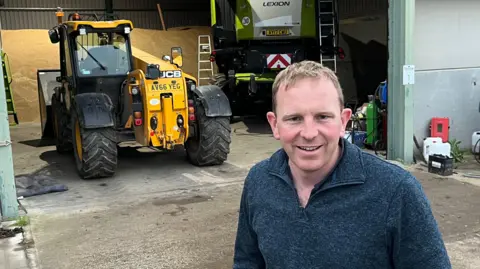 JOZEF HALL/BBC Tom Mead smiles and looks directly at the camera as he is photographed outside in front of a yellow tractor. He is wearing a blue jumper and dark trousers and has both hands in his pockets. Behind him is a large barn with farmyard items on the ground behind. 