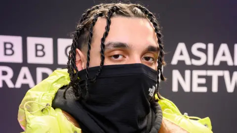 Tracey Welch/ BBC Sliime in the BBC Asian Network studio in February 2024. The rapper wears a bright green puffer jacket and covers the bottom half of his face with a black scarf. 