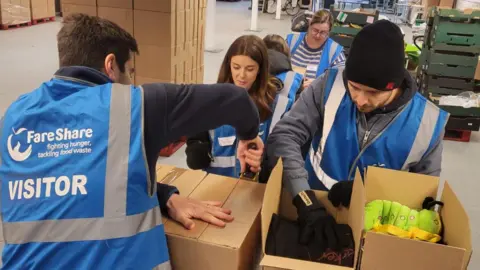 BBC Five people are in a warehouse packing cardboard boxes. They are wearing blue high-vis vests. There are two men in the foreground and a woman who is smiling.