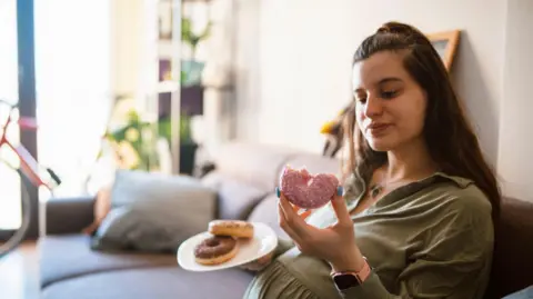 Getty Images Pregnant woman sits on a sofa holding a plate of doughnuts with pink and chocolate-toppings. The pink doughnut is half-eaten.